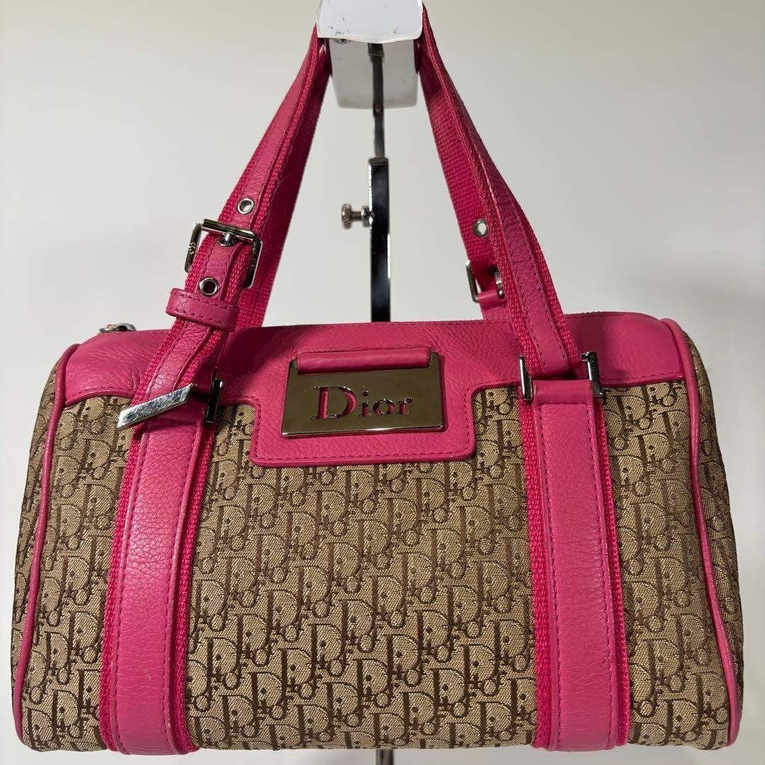 A Dior satchel handbag with a beige and brown monogram canvas body, bright pink leather trim and handles, and a gold-tone Dior logo plaque on the front, displayed on a stand against a plain light background.