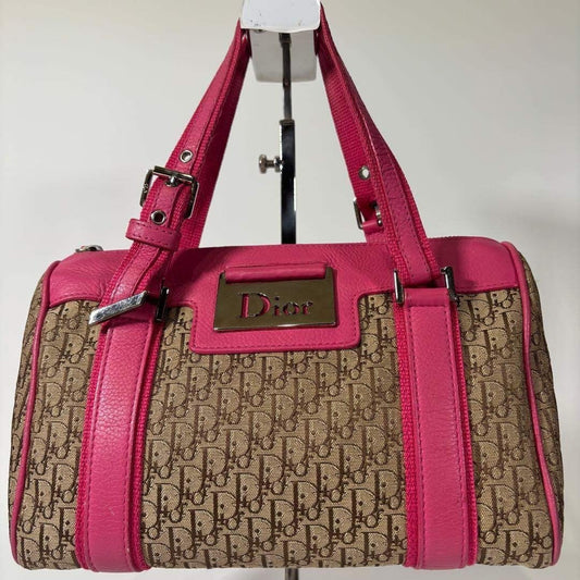 A Dior satchel handbag with a beige and brown monogram canvas body, bright pink leather trim and handles, and a gold-tone Dior logo plaque on the front, displayed on a stand against a plain light background.