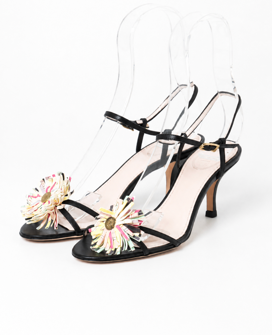 A pair of Pucci black strappy kitten heeled sandals with clear acrylic heels and colorful fringe flower embellishments on the toes, displayed on clear acrylic stands against a white background.